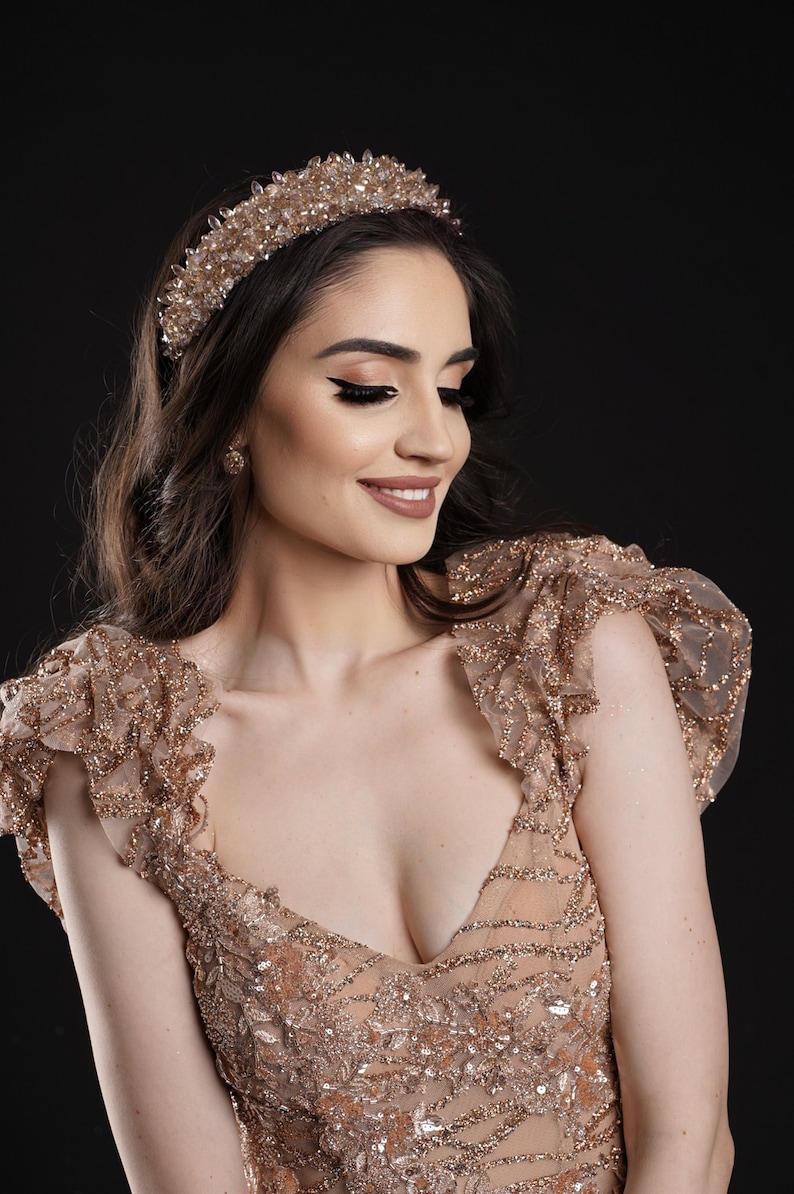 May include: A woman wearing a gold-colored, beaded tiara and a matching gown with a plunging neckline and ruffled sleeves. The dress and headpiece are adorned with sparkling embellishments, set against a dark background.