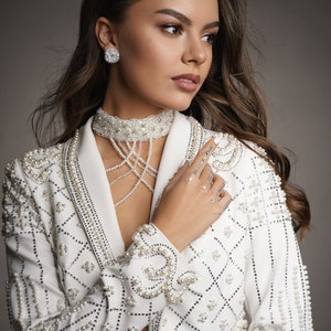 Op de afbeelding: Een vrouw draagt een witte blazer met parelversiering. De blazer heeft een geometrisch patroon en is versierd met parels. Ze draagt een parelketting en oorbellen.