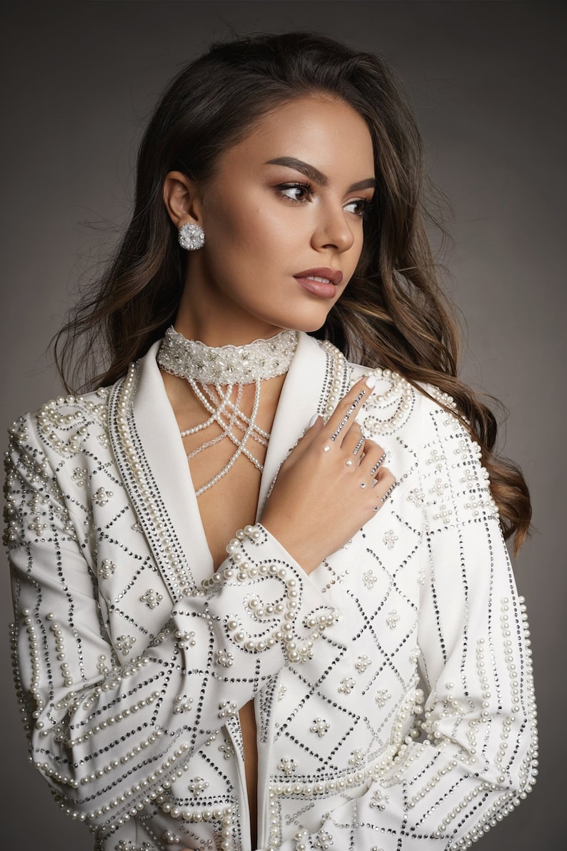 Op de afbeelding: Een vrouw draagt een witte blazer met parelversiering. De blazer heeft een geometrisch patroon en is versierd met parels. Ze draagt een parelketting en oorbellen.