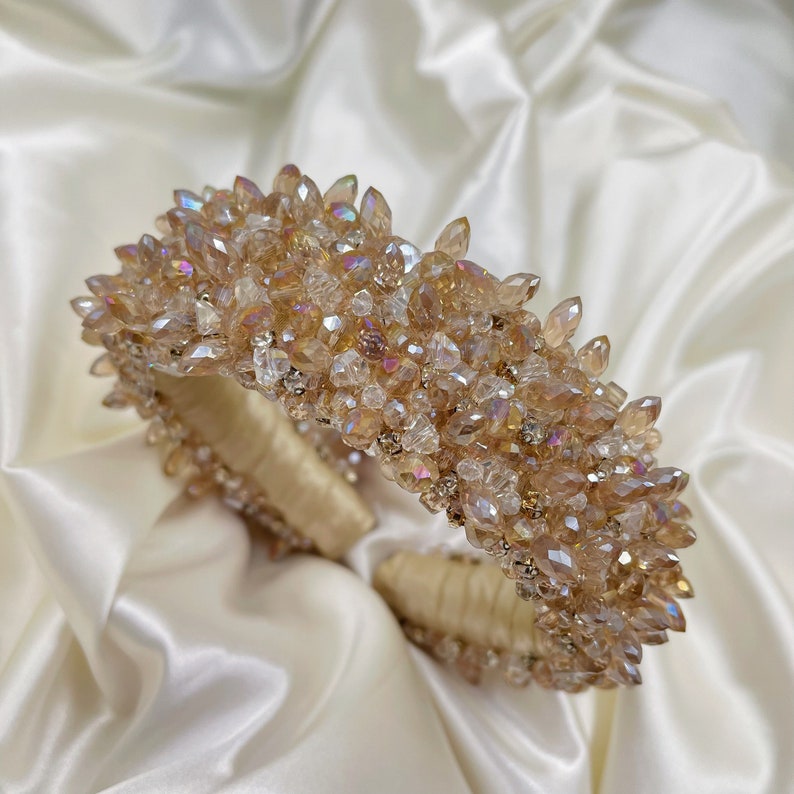 May include: A gold-toned headband, densely adorned with sparkling, faceted crystals. The crystals vary in size and shape, creating a textured, eye-catching surface. The band itself is wrapped in a soft, cream-colored fabric.