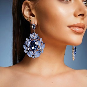 May include: A pair of large, statement earrings with a blue and white crystal design. The earrings feature a large, oval-shaped blue crystal surrounded by smaller, teardrop-shaped white crystals.