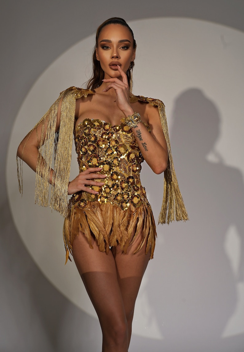 Puede incluir: Una mujer con un traje dorado con lentejuelas y flecos, con un corpi&ntilde;o sin tirantes y una falda con ribete de plumas. El traje tiene un ribete de flecos dorados en los hombros. La mujer tiene un tatuaje en el brazo que dice "Hope Love".