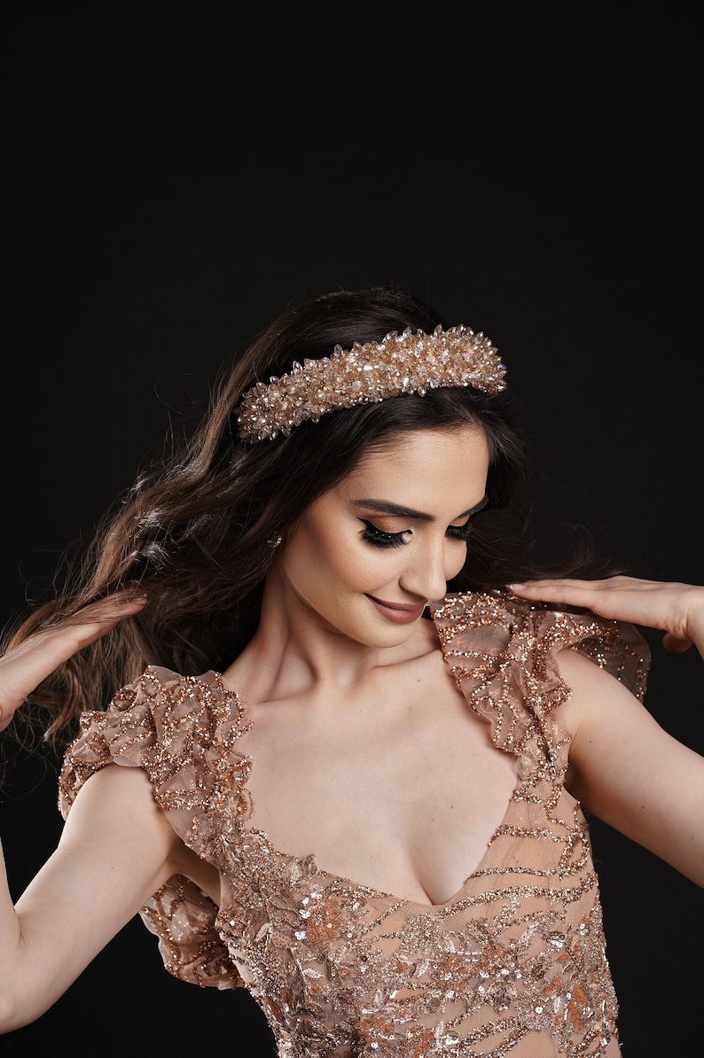 May include: A gold-toned crystal-studded headband. The headband is the focal point, set against a dark background. The model is wearing a matching gold-toned, embellished dress with a low neckline and ruffled sleeves. The overall aesthetic is elegant and glamorous.