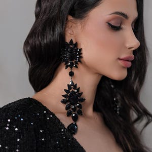 Peut inclure: Une femme aux longs cheveux noirs portant une robe noire à paillettes et de grandes boucles d'oreilles en cristal noir.