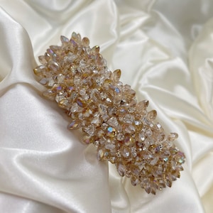 May include: A decorative hair clip featuring a cluster of iridescent, champagne-colored crystals and small clear rhinestones. The clip is rectangular and set against a white satin background, showcasing its sparkling details.