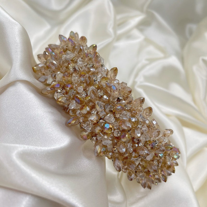 May include: A decorative hair clip featuring a cluster of iridescent, champagne-colored crystals and small clear rhinestones. The clip is rectangular and set against a white satin background, showcasing its sparkling details.
