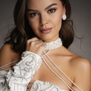 Op de afbeelding: Een vrouw draagt een wit, off-shoulder topje met parelversieringen. Het topje heeft een parelketting en een parelketting die over &eacute;&eacute;n schouder gedrapeerd is.