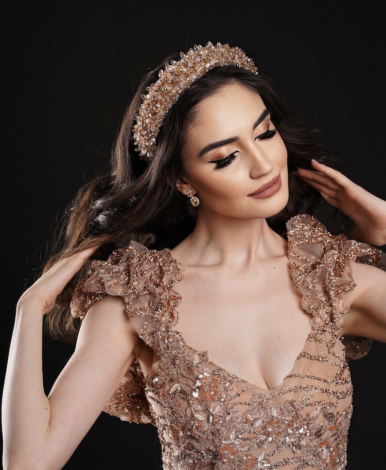 May include: A gold-toned bridal crown with clear crystal accents. The crown sits atop long, wavy brown hair. The model is wearing a matching gold-toned dress with a plunging neckline and embellished with sequins and beads. The background is black.