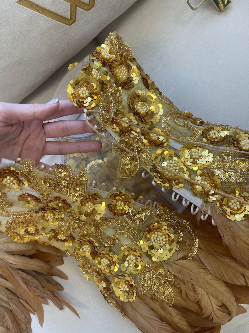 Puede incluir: Un top dorado con lentejuelas y cuentas con plumas marrones. El top est&aacute; hecho de una tela transparente y tiene un dise&ntilde;o floral.
