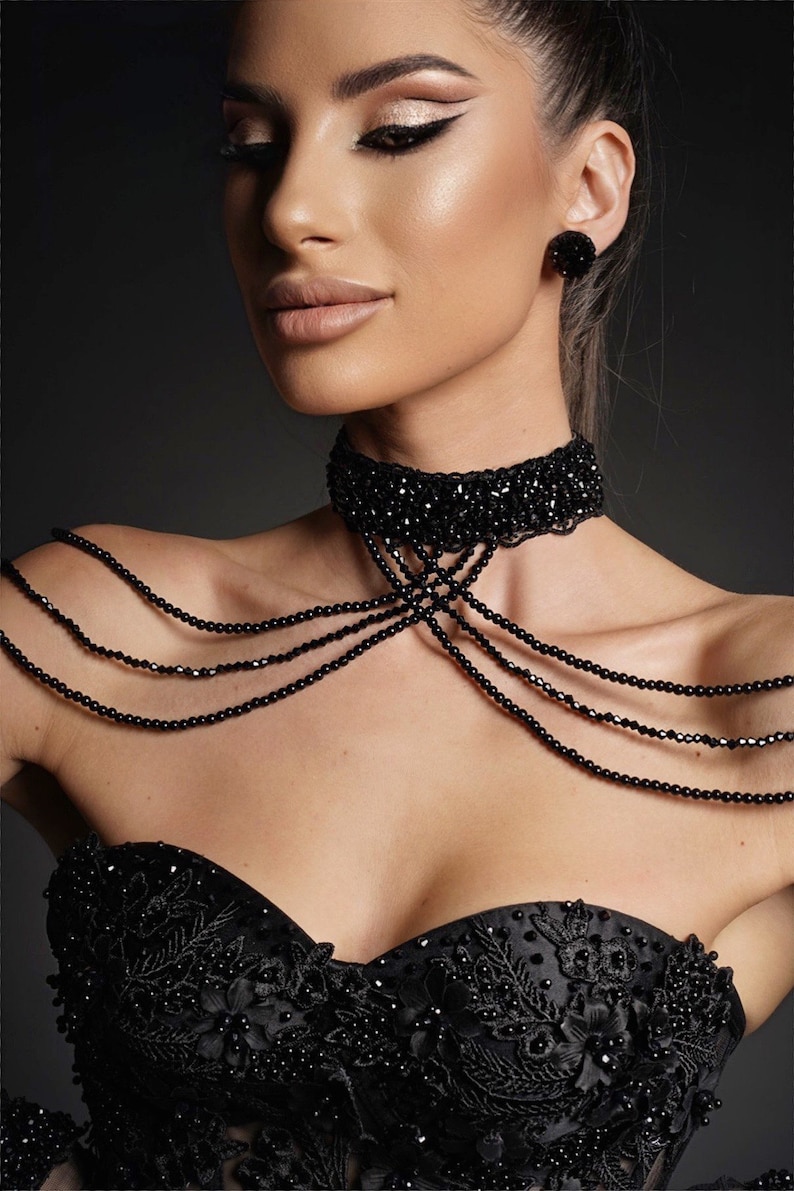 Op de afbeelding: Een vrouw draagt een zwarte strapless jurk met parelversieringen. Ze draagt een zwarte choker ketting en bijpassende oorbellen.