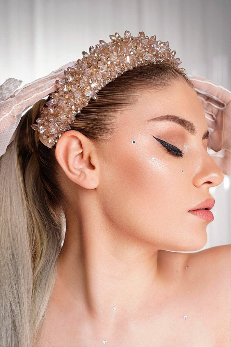 May include: A side profile of a person wearing a sparkling, champagne-colored crystal crown. The crown is adorned with numerous crystals. The person has long blonde hair and is wearing makeup, including black eyeliner and small face gems.