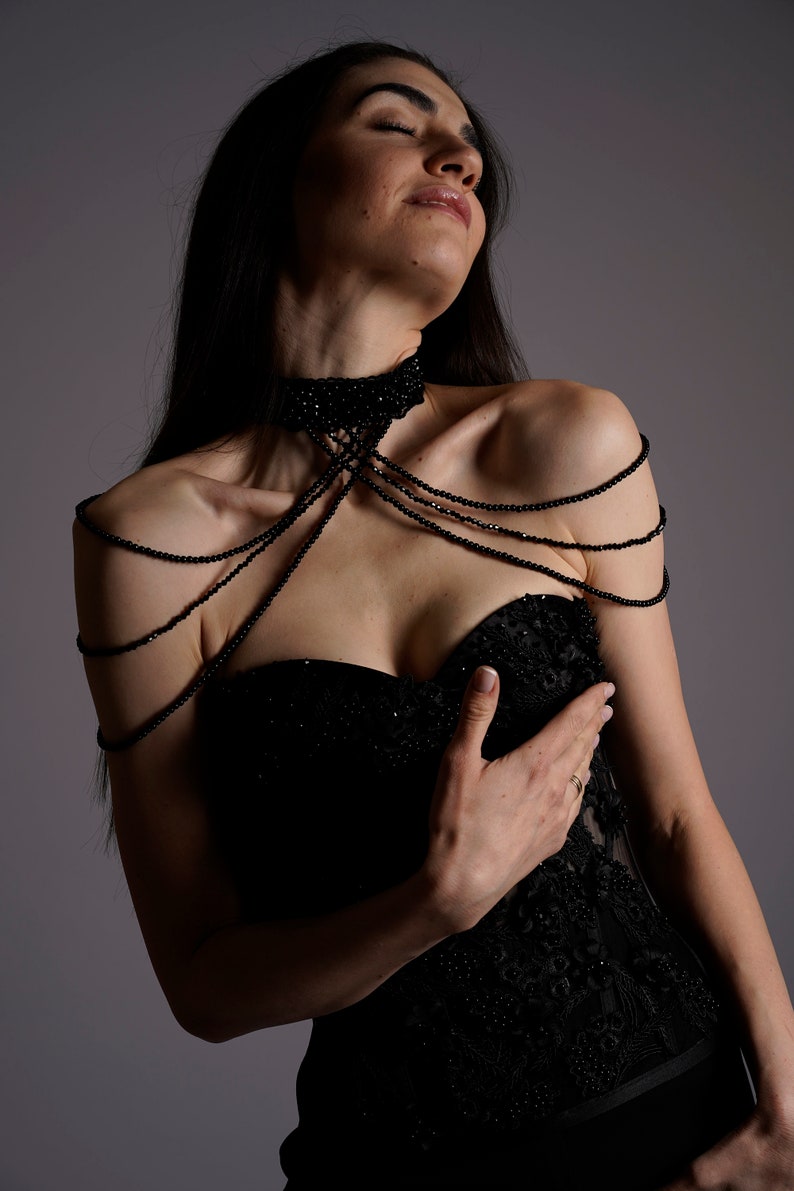 Op de afbeelding: Een vrouw die een zwarte strapless jurk draagt met een kralenhalsband en bijpassende kralen schouderbandjes. De jurk heeft een kant overlay en is strak gesneden.