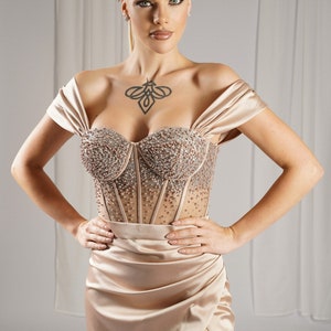 May include: A formal, champagne-colored gown with a beaded bodice and a high slit. The off-the-shoulder dress features a sweetheart neckline and is paired with a jeweled headband. The model has blonde hair and a tattoo on their chest.