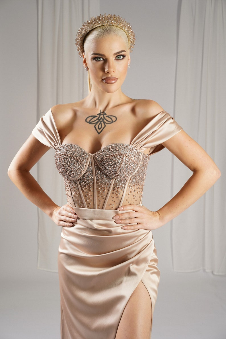 May include: A formal, champagne-colored gown with a beaded bodice and a high slit. The off-the-shoulder dress features a sweetheart neckline and is paired with a jeweled headband. The model has blonde hair and a tattoo on their chest.