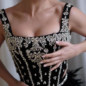 May include: Black evening gown with a square neckline and embellished with sparkling crystals. The bodice features intricate floral and linear crystal designs. The skirt is trimmed with black feathers, creating a glamorous look.