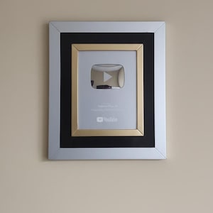 以下が含まれることがあります： 額縁に入ったYouTube Play Buttonアワード。アワードは、シルバーのフレーム、黒い内側のボーダー、金色の内側のフレームを備えています。中央には、シルバーの再生ボタンのロゴとYouTubeのロゴがあります。