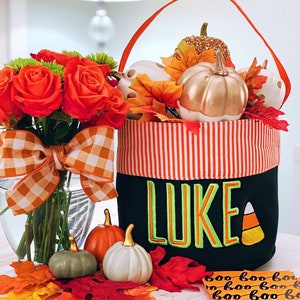 May include: A black fabric bucket with an orange handle and a striped orange and white top band. The name LUKE is embroidered in green letters on the front, next to a candy corn graphic. It is filled with small pumpkins and autumn leaves.