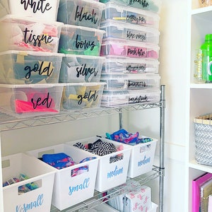 May include: A white metal shelf unit with clear plastic storage bins labeled with black marker. The bins are filled with various craft supplies, including tissue paper, vinyl, and ribbon. The shelf unit is in a white closet with a gray and white patterned basket on the shelf to the right.