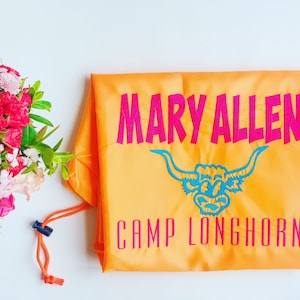 May include: Orange drawstring bag with the text "MARY ALLEN CAMP LONGHORN" in pink lettering. A blue embroidered longhorn bull head is centered above the text.