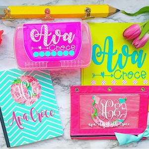 May include: A set of personalized school supplies featuring a turquoise and white chevron pattern with the name "Ava Croce" and a floral monogram. The set includes a clear pencil box, a binder, and a clear pencil pouch with a pink background.