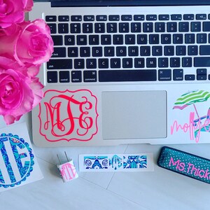 Könnte beinhalten: Ein Laptop mit mehreren Aufklebern auf dem Deckel und dem Trackpad. Die Aufkleber beinhalten ein Monogramm, eine Strandszene mit einem Sonnenschirm und Stühlen sowie den Text "Molly Jean". Außerdem gibt es einen blaugrünen Hefter mit dem Text "Ms. Thicksten" darauf.