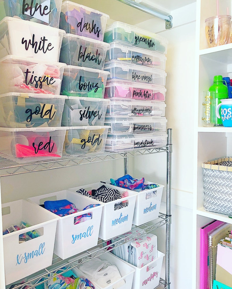 May include: A white metal shelf unit with clear plastic storage bins labeled with black marker. The bins are filled with various craft supplies, including tissue paper, vinyl, and ribbon. The shelf unit is in a white closet with a gray and white patterned basket on the shelf to the right.