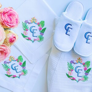 May include: Two white bath slippers with embroidered floral designs and the initials "CE" in blue thread. Two white linen napkins with the same floral design and initials are also included.