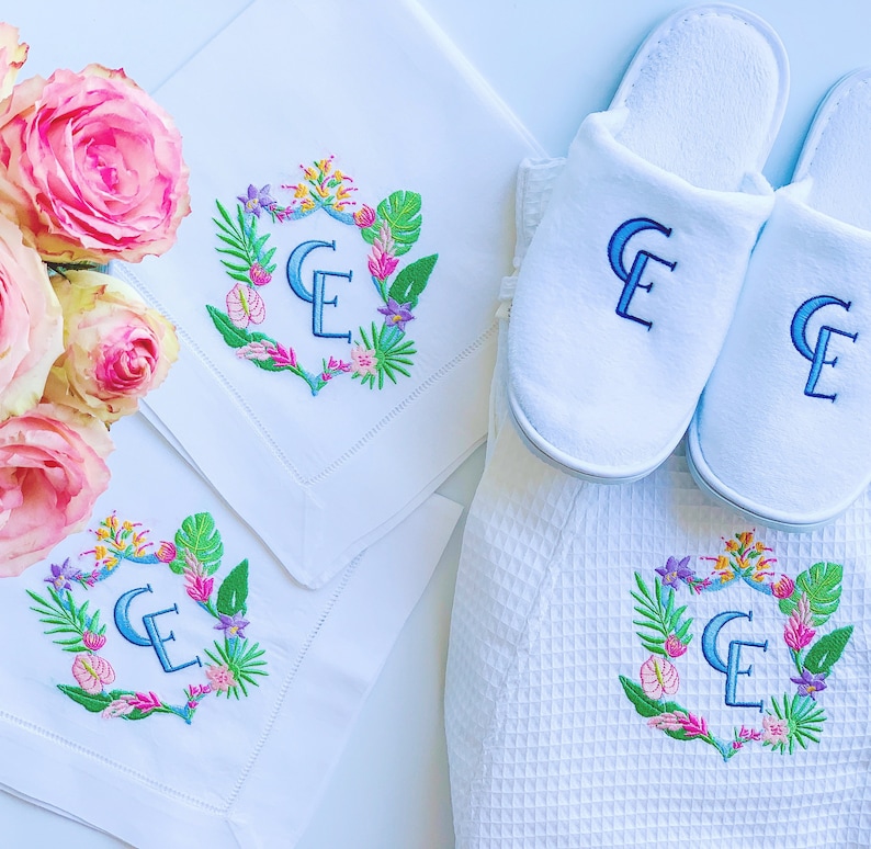 May include: Two white bath slippers with embroidered floral designs and the initials "CE" in blue thread. Two white linen napkins with the same floral design and initials are also included.