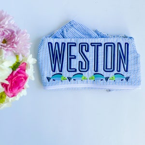 May include: A blue and white striped fabric with the name "WESTON" embroidered in navy blue. A row of small green and blue fish are embroidered below the name.