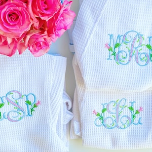 May include: Two white waffle knit robes with embroidered monograms. The monograms are in blue with green leaves and pink flowers. The monograms are "CSR" and "MRL".