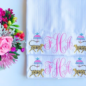 May include: Two white pillowcases with pink embroidered monograms and a leopard print design with a colorful umbrella. The pillowcases have a subtle striped pattern.
