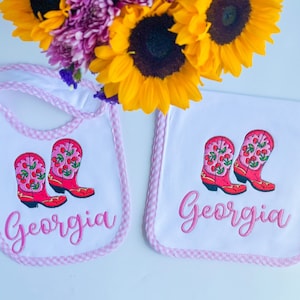 May include: Two white burp cloths with pink trim and embroidered red cowboy boots with cherries. The text "Georgia" is embroidered on each cloth.