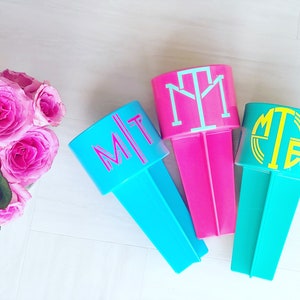 May include: Three personalized drink holders with different monogram designs. The holders are turquoise, pink, and teal. The monograms are in white, yellow, and pink.