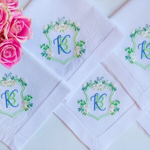 May include: Four white linen napkins with embroidered green and blue floral designs and the letter "K" in a shield shape.