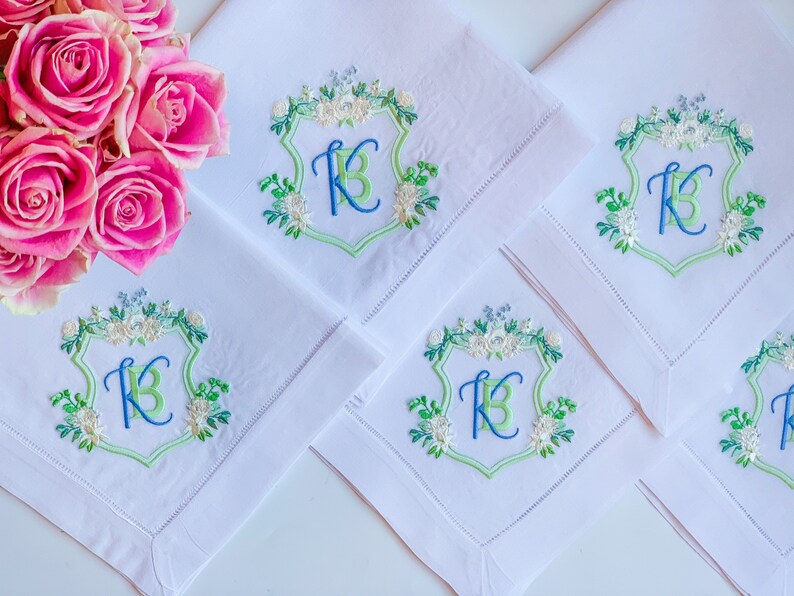 May include: Four white linen napkins with embroidered green and blue floral designs and the letter "K" in a shield shape.