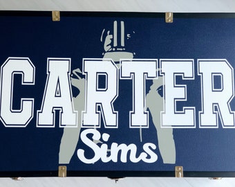 May include: A navy blue wooden box with a white silhouette of a person wearing a helmet and the name "Carter Sims" in white letters.