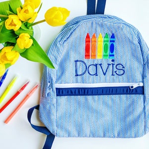 May include: A blue and white striped backpack with a zipped pocket and a rainbow of crayons embroidered on the front. The name "Davis" is also embroidered on the front of the backpack.