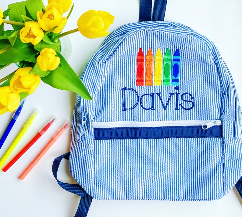 May include: A blue and white striped backpack with a zipped pocket and a rainbow of crayons embroidered on the front. The name "Davis" is also embroidered on the front of the backpack.