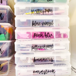 May include: Clear plastic storage bins stacked on a metal shelf. Each bin is labeled with black text: green htv, wrapping paper, blue vinyl, pink htv, patterned vinyl, banners, cardstock.