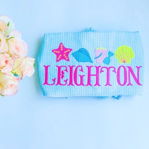May include: A light blue personalized  towel with the name Leighton embroidered in pink. The towel features a starfish, seashells, and a bucket and shovel in bright colors.