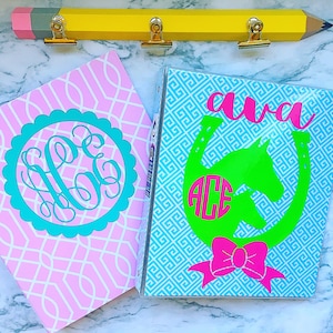 May include: Two personalized binders with decorative designs. One is pink with a teal monogram, the other is blue with a horse and horseshoe design, the name "ava", and a pink bow. A yellow pencil-shaped clip holder is above.