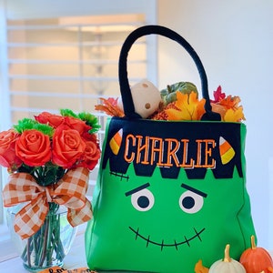 May include: A bright green tote bag featuring a black top border with the embroidered name CHARLIE in orange, flanked by candy corn icons. The bag has a stitched Frankenstein face with black eyes and a mouth, plus two black fabric handles.