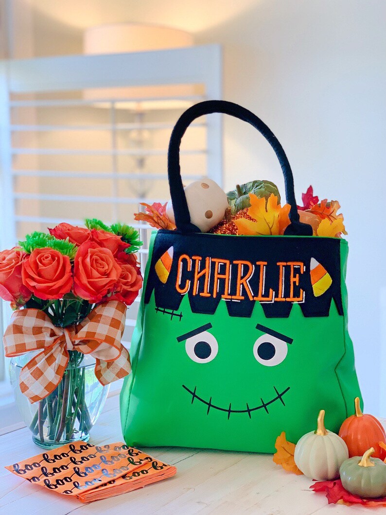 May include: A bright green tote bag featuring a black top border with the embroidered name CHARLIE in orange, flanked by candy corn icons. The bag has a stitched Frankenstein face with black eyes and a mouth, plus two black fabric handles.