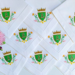 May include: Eight white linen napkins with embroidered green shields featuring a gold crown and the letter "A" in red. Each shield is surrounded by yellow, pink, and green floral embroidery.