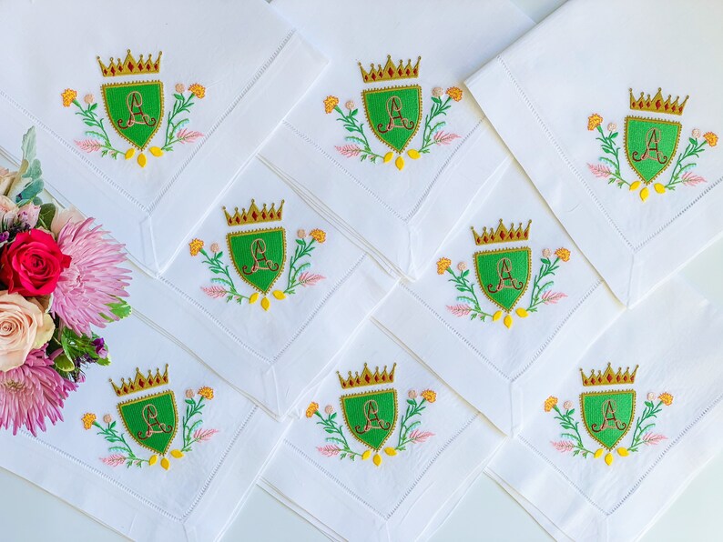 May include: Eight white linen napkins with embroidered green shields featuring a gold crown and the letter "A" in red. Each shield is surrounded by yellow, pink, and green floral embroidery.