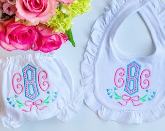 Puede incluir: Juego de babero y braguita de bebé blancos con monograma bordado en rosa y azul. El babero y la braguita tienen un borde con volantes y un diseño floral. Un ramo de rosas rosas y flores verdes está en el fondo.