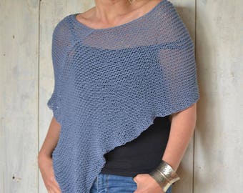 Puede incluir: Un poncho azul de punto con una trama suelta y abierta. El poncho está drapeado sobre una persona que lleva una blusa negra y unos vaqueros azules.