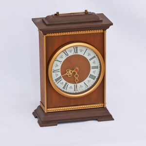 May include: A brown wooden mantel clock with a gold-colored face and Roman numeral hour markers. The clock has a decorative gold trim around the face and a gold-colored base.