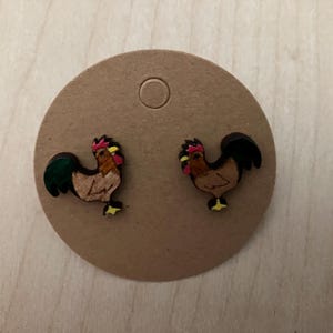 May include: A pair of wooden rooster stud earrings. Each earring features a brown rooster with a red comb, yellow beak, and black tail feathers. The earrings are displayed on a round, tan earring card.