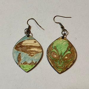 May include: A pair of wooden earrings with a green alien face on one and a flying saucer with a person standing in front of it on the other.